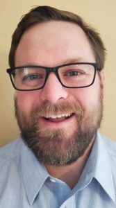 Portrait photo of Joseph Landry, Logistics Specialist - DTS Logistics. Joseph is smiling amiably, wearing a light blue button down oxford dress shirt, and has thick-rimmed glasses and a full beard.